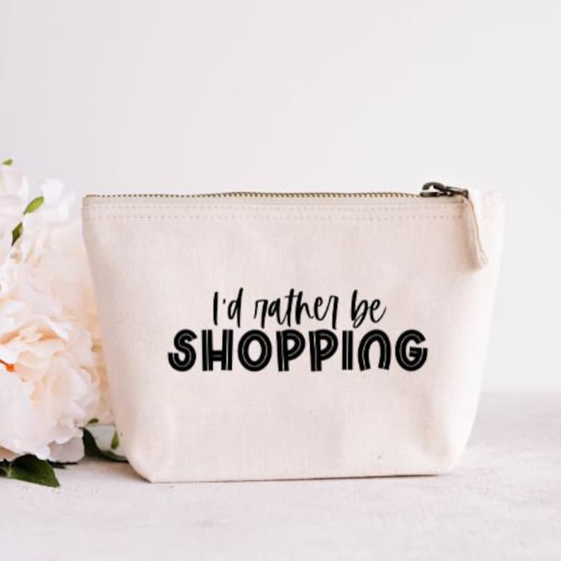 May include: A cream-colored canvas cosmetic bag with a zipper closure. The bag features the phrase "I'd rather be SHOPPING" in bold black letters. A bouquet of white flowers is visible in the background.