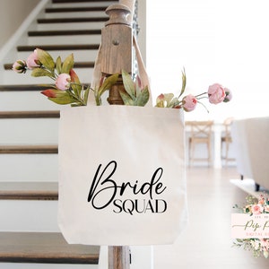Op de afbeelding: Een cr&egrave;mekleurige draagtas met de woorden "Bride Squad" in zwart script. De tas is gevuld met roze en groene kunstbloemen. De tas hangt aan een houten balustrade. De achtergrond omvat een trap en een woonkamer.