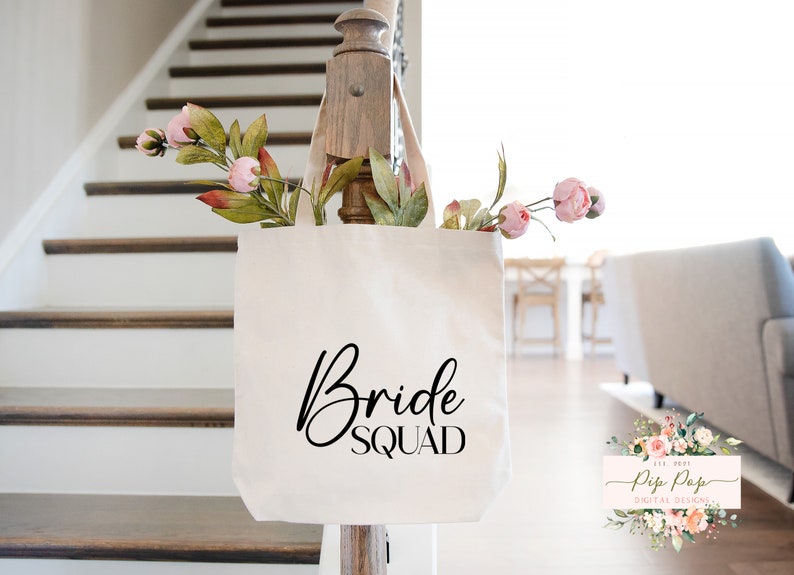 Op de afbeelding: Een cr&egrave;mekleurige draagtas met de woorden "Bride Squad" in zwart script. De tas is gevuld met roze en groene kunstbloemen. De tas hangt aan een houten balustrade. De achtergrond omvat een trap en een woonkamer.