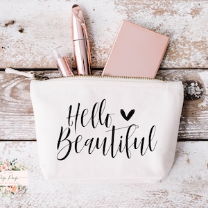 Peut inclure: Une pochette zippée en toile blanche avec le texte "Hello Beautiful" en écriture noire avec un petit cœur noir. La pochette est partiellement ouverte et contient un miroir compact rose gold, un tube de mascara rose gold et un tube transparent de brillant à lèvres.