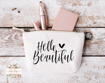May include: White canvas zippered pouch with the text "Hello Beautiful" in black script with a small black heart. The pouch is partially open and contains a rose gold compact mirror, a rose gold mascara tube, and a clear tube of lip gloss.
