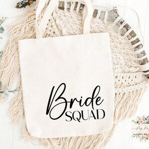 Op de afbeelding: Cr&egrave;mekleurige draagtas met de woorden "Bride Squad" in zwart script. De tas heeft twee handvatten en is gemaakt van een stevige stof. De tas ligt op een wit oppervlak met decoratieve elementen.