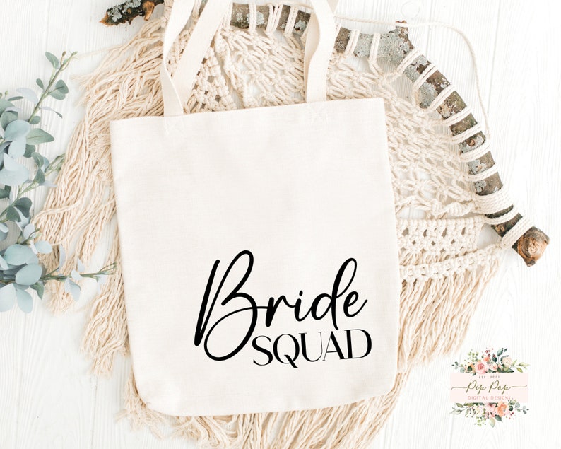 Op de afbeelding: Cr&egrave;mekleurige draagtas met de woorden "Bride Squad" in zwart script. De tas heeft twee handvatten en is gemaakt van een stevige stof. De tas ligt op een wit oppervlak met decoratieve elementen.