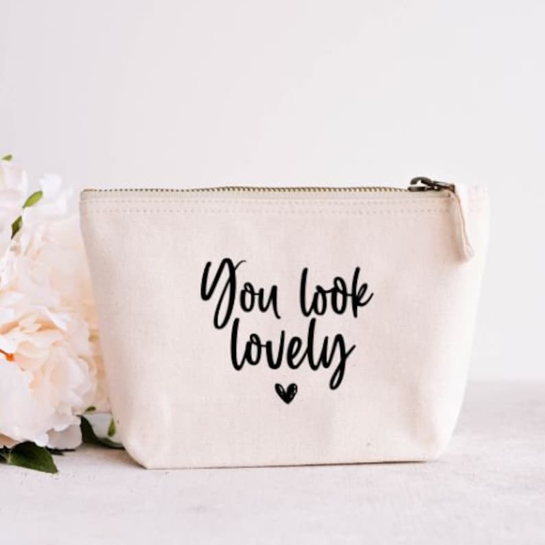 May include: A cream-colored canvas cosmetic bag with a zipper closure. The bag features the phrase "You look lovely" in black script with a small heart below. Soft, white flowers are visible in the background.