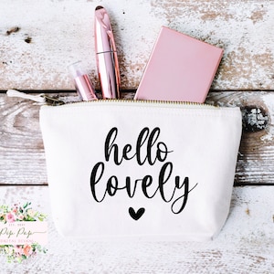 May include: White canvas zippered pouch with the text "hello lovely" in black script with a small black heart below. The pouch is partially open and contains a pink compact, a rose gold lipstick, and a rose gold mascara.