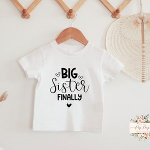 Peut inclure: T-shirt blanc avec un texte noir qui dit "Big Sister Finally" avec un petit cœur au-dessus et en dessous du texte.