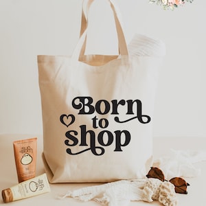 May include: A white canvas tote bag with black text that says "Born to Shop" with a heart symbol. The bag is sitting on a white surface with two bottles of sunscreen and a pair of sunglasses.