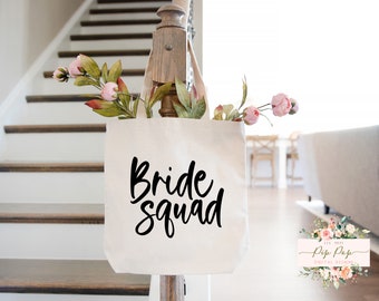 Op de afbeelding: Witte canvas tote bag met zwarte tekst die "Bride Squad" zegt. De tas hangt aan een houten leuning met roze bloemen op de achtergrond.