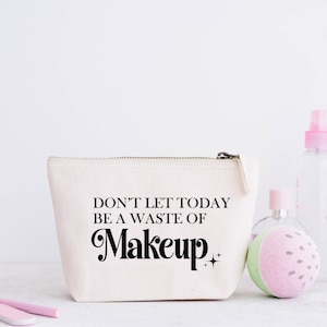 May include: A cream-colored canvas makeup bag with a zipper closure. The bag features the text "DON'T LET TODAY BE A WASTE OF Makeup" in black script. Other beauty products are in the background.