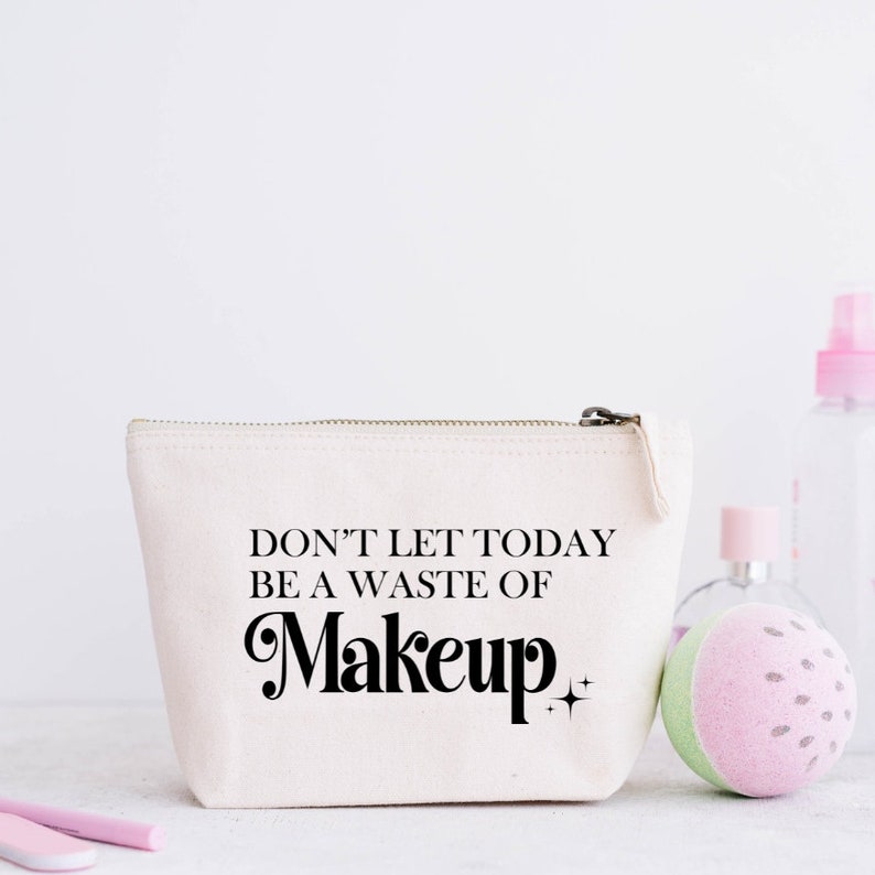 May include: A cream-colored canvas makeup bag with a zipper closure. The bag features the text "DON'T LET TODAY BE A WASTE OF Makeup" in black script. Other beauty products are in the background.