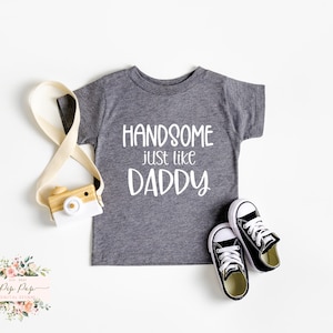 Puede incluir: Una camiseta gris de manga corta con el texto "HANDSOME just like DADDY" en blanco. La camiseta está sobre un fondo blanco con una cámara de juguete de madera y un par de zapatillas negras.