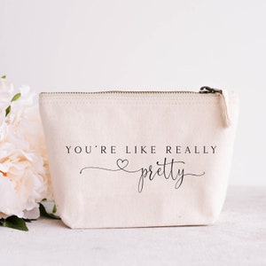 May include: Cream-colored canvas cosmetic bag with a zipper closure. The bag features the text "YOU'RE LIKE REALLY pretty" in black script. A bouquet of light pink flowers is in the background.