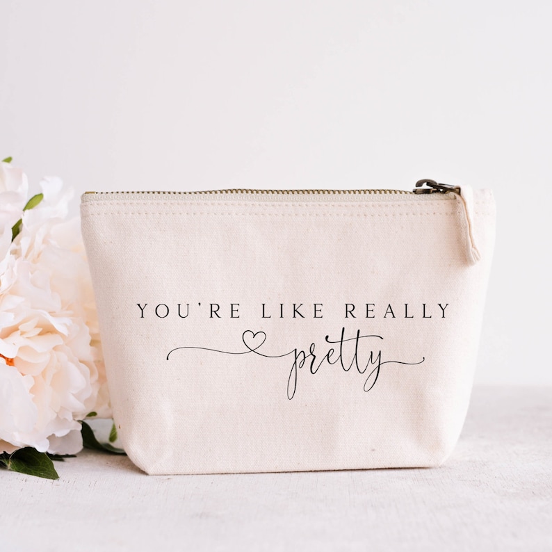 May include: Cream-colored canvas cosmetic bag with a zipper closure. The bag features the text "YOU'RE LIKE REALLY pretty" in black script. A bouquet of light pink flowers is in the background.