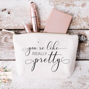 May include: White canvas zippered pouch with the text "you're like really pretty" in black cursive lettering. The pouch is partially open and contains a pink compact, a clear tube of lipstick, and a gold mascara tube.