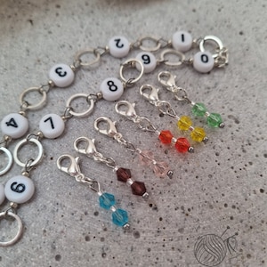 Könnte beinhalten: Strickmaschenmarkierer und Reihenzähler. Die Reihenzähler sind weiße Perlen mit schwarzen Zahlen. Die Maschenmarkierer sind silberfarben mit farbigen Perlen in Blau, Braun, Rosa, Rot, Gelb und Grün.