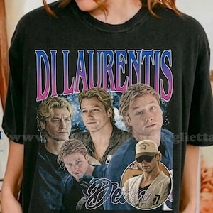 Può includere: T-shirt nera con una grafica di più immagini di una persona con il testo "DI LAURENTIS" in un font rosa e blu. La parola "Dean" è scritta in corsivo sotto le immagini. La maglietta è in cotone pesante.