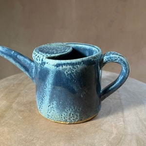 May include: A small, blue ceramic jug with a lid and a handle. The jug has a speckled glaze and a gold rim.