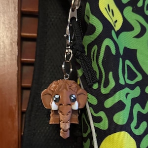 Puede incluir: Un llavero de mamut de dibujos animados marrón y blanco con ojos grandes y colmillos. El llavero está unido a un tirador de cremallera plateado en una mochila negra y verde.