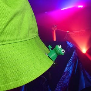 Puede incluir: Un sombrero de pescador verde brillante con una pequeña figura de rana verde sujeta al borde. El sombrero tiene un borde cosido y un pequeño detalle puntiagudo en la parte superior. El fondo es oscuro con iluminación de escenario roja y azul.