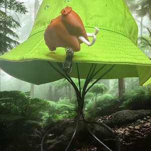 May include: A bright green bucket hat with a brown turkey-shaped clip attached. The hat is displayed on a metal stand with a circular base and tree-like support. The background is a blurred forest scene.