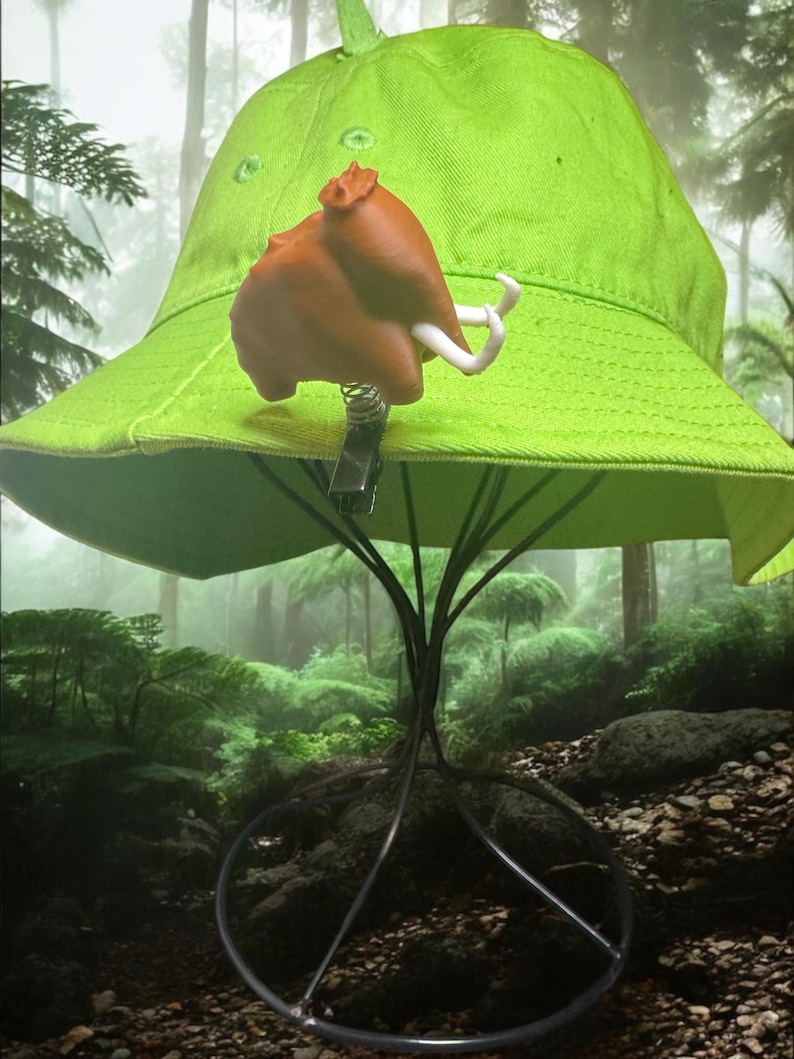 May include: A bright green bucket hat with a brown turkey-shaped clip attached. The hat is displayed on a metal stand with a circular base and tree-like support. The background is a blurred forest scene.
