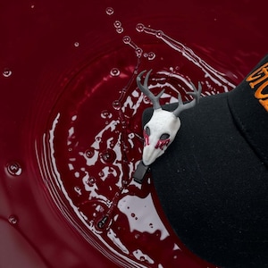 May include: A white deer skull pin with grey antlers and red accents, attached to a black hat. The hat has orange text on the side. The background is a deep red liquid with bubbles.