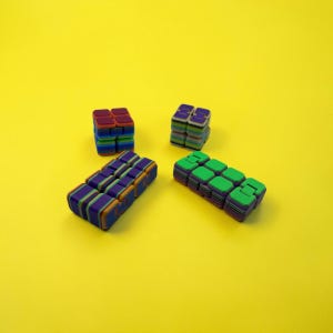 May include: Four colorful fidget toys on a yellow background. The toys are in various shapes, including cubes and rectangular bars. The colors include red, blue, green, purple, and orange. These toys are designed to help with focus and stress relief.