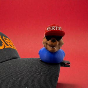 May include: A black baseball cap with a clip, featuring a brown bear figurine wearing a red cap with "GRIZ" in white letters and sunglasses. The bear is wearing a blue shirt, set against a red background.
