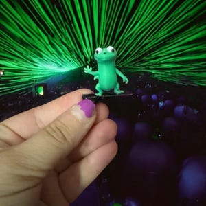 May include: A small, green frog figurine with large white eyes and outstretched arm. The frog is standing on a small, dark platform. The background features a vibrant display of green laser beams.