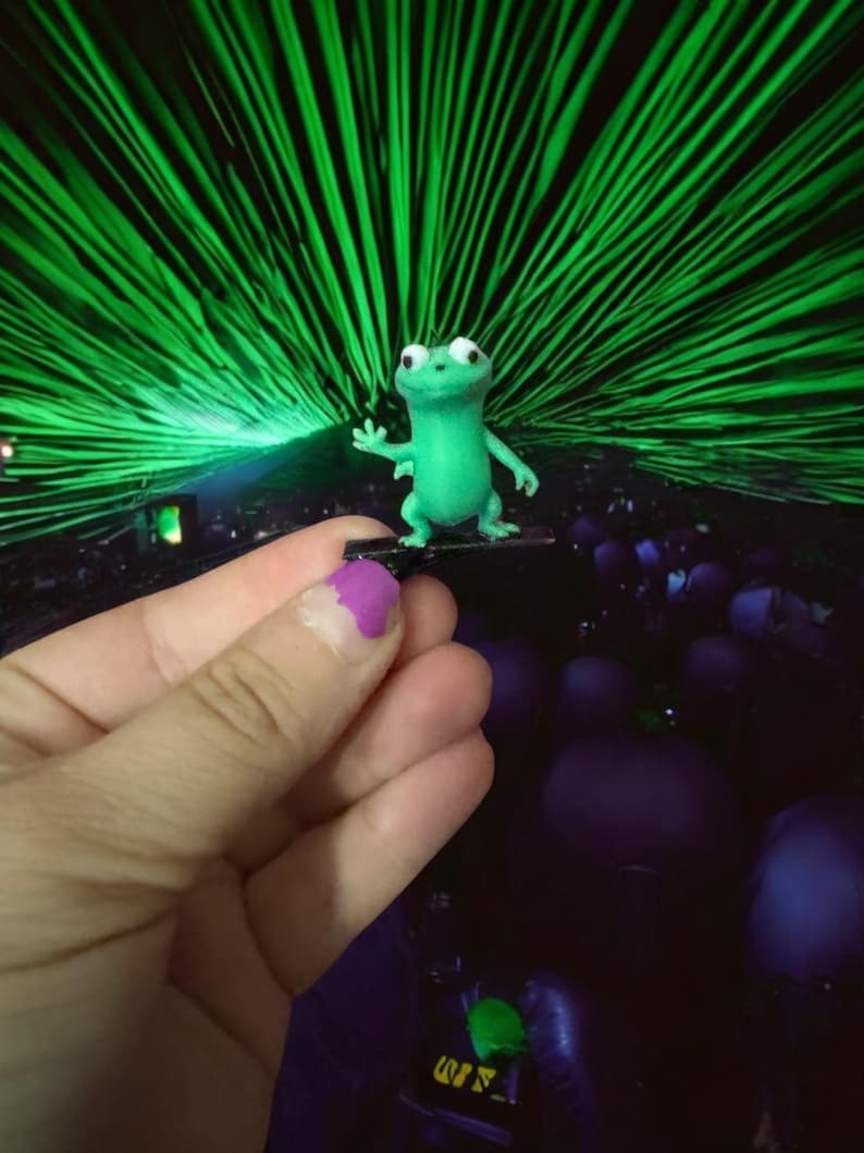 May include: A small, green frog figurine with large white eyes and outstretched arm. The frog is standing on a small, dark platform. The background features a vibrant display of green laser beams.