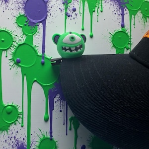 Puede incluir: Un clip de sombrero verde con forma de monstruo con un solo ojo y una amplia sonrisa, sujeto a una gorra de béisbol negra. El fondo es blanco con salpicaduras y goteos de pintura verde y morada.