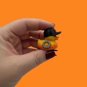 Puede incluir: Un pato de goma amarillo con una gorra de béisbol negra con un logotipo rojo y un diseño de engranaje plateado. El pato tiene llamas naranjas alrededor de su cuerpo. El fondo es de color naranja.