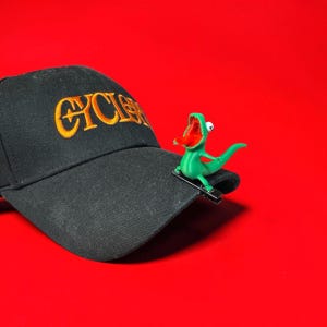 May include: A black baseball cap featuring the embroidered yellow text CYCLOPS on the front panel. A small, bright green lizard figure with an open red mouth and a single white eye is attached to the brim of the cap with a black metal clip.