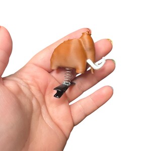 May include: A brown mammoth-shaped clip with white tusks, a black clip, and a silver spring. The clip is held in a hand against a white background. The mammoth is a light brown color.