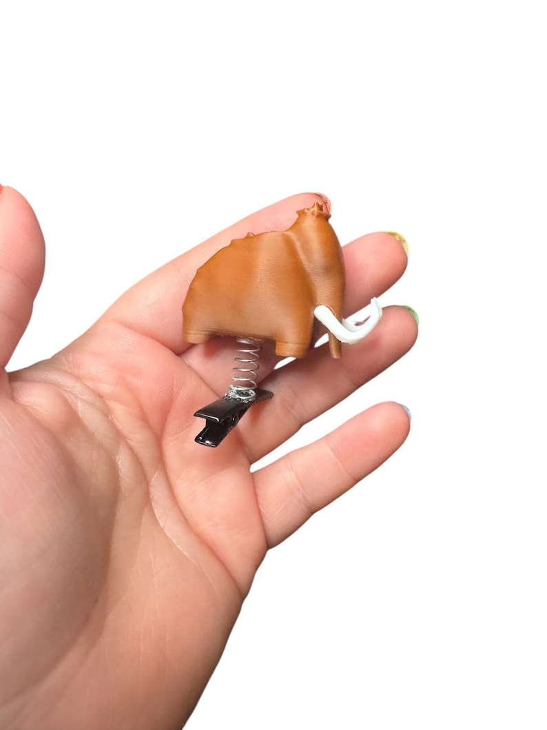 May include: A brown mammoth-shaped clip with white tusks, a black clip, and a silver spring. The clip is held in a hand against a white background. The mammoth is a light brown color.