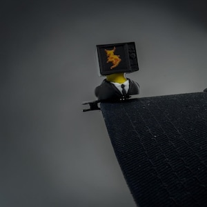 May include: A black hat clip accessory featuring a figure with a television set for a head. The screen displays a yellow creature graphic. The figure wears a black suit jacket with a white shirt and a black tie, attached to a black brimmed hat.