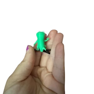 May include: A small, bright green frog figurine with white eyes and outstretched arms. The frog is a solid color and appears to be made of plastic. The figurine is held in a person's hand against a white background.