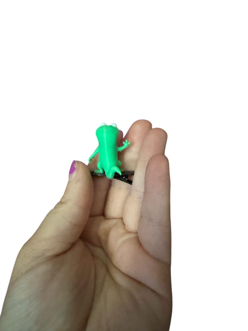 May include: A small, bright green frog figurine with white eyes and outstretched arms. The frog is a solid color and appears to be made of plastic. The figurine is held in a person's hand against a white background.