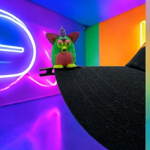 May include: A green, pink, and yellow Furby toy sits on the peak of a black baseball cap. The Furby has large eyes and a small beak. The background features neon lights and colourful walls.