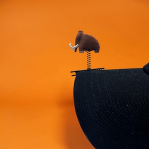 May include: A brown woolly mammoth figurine with white tusks attached to a thin metal spring, which is mounted onto a black alligator clip. The clip is secured to the edge of a black baseball cap brim, creating a bobblehead effect.