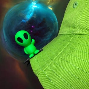 May include: A lime green baseball cap with a small, green alien figurine attached. The alien has large black eyes and is waving. The background features a nebula with blue and yellow hues.