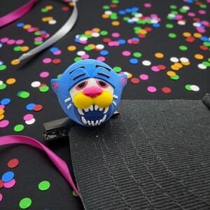 May include: A blue, 3D-printed tiger face hair clip with a white, yellow, and pink face. The clip is attached to a black fabric surface, with colourful confetti and ribbons in the background.