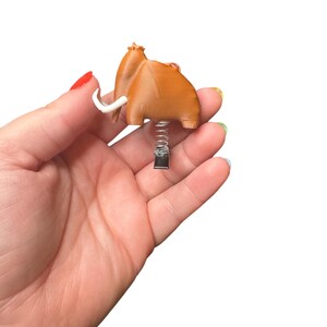 May include: A brown, cartoon-style mammoth figurine with white tusks and a spring base. The figurine is held in a hand against a white background. The mammoth has a textured appearance.