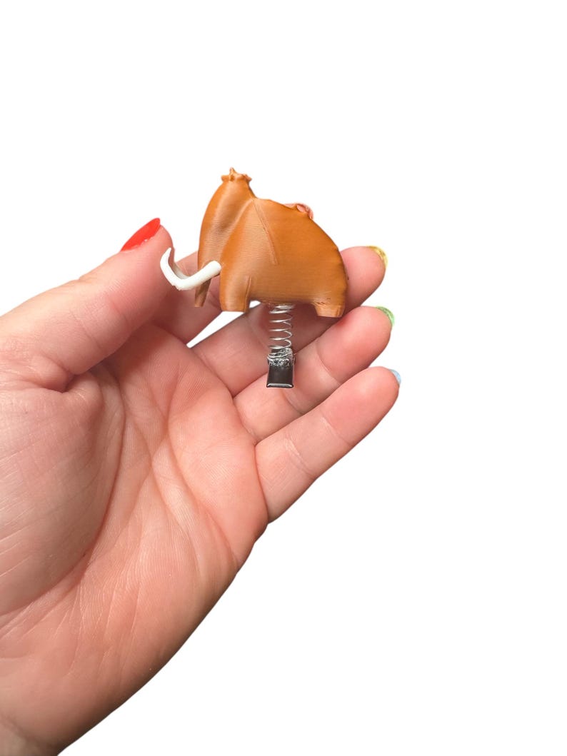May include: A brown, cartoon-style mammoth figurine with white tusks and a spring base. The figurine is held in a hand against a white background. The mammoth has a textured appearance.