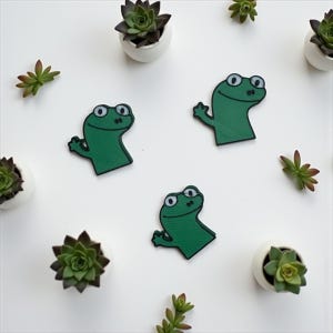 以下が含まれることがあります： 白い背景に、白い鉢植えの多肉植物と並んで配置された、白と青の目を持つ緑色のカエル型のパッチのコレクション。 パッチは笑顔で、腕を広げています。