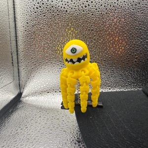 May include: A yellow, cartoonish octopus-like creature with one eye and a toothy grin. The tentacles are segmented, and the eye has a green iris. The figure is set against a textured, silver background.