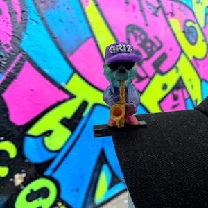 Könnte beinhalten: Eine kleine, 3D-gedruckte Figur eines türkisfarbenen Bären mit einer violetten Kappe mit der Aufschrift GRIZ, einer schwarzen Sonnenbrille und einer violetten Jacke. Der Bär hält ein gelbes Saxophon und ist an einem schwarzen Clip am Schirm befestigt.