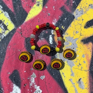Puede incluir: Una pulsera de cuentas rojas, amarillas y negras con un dije circular central amarillo que muestra el texto ALL DAT en rojo. Cuatro dijes circulares amarillos a juego con el mismo texto rojo están dispuestos alrededor de la pulsera.