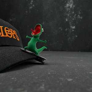May include: A green lizard figurine with a red mouth and tongue is attached to a black hat. The hat has the word "LERS" embroidered in orange. The lizard is on a small black skateboard.