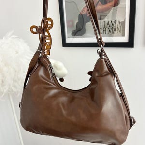 May include: A brown hobo-style shoulder bag with a smooth finish, featuring a top zipper closure and long adjustable straps. A translucent amber-colored claw clip is attached to one of the shoulder straps, adding a decorative element to the bag.